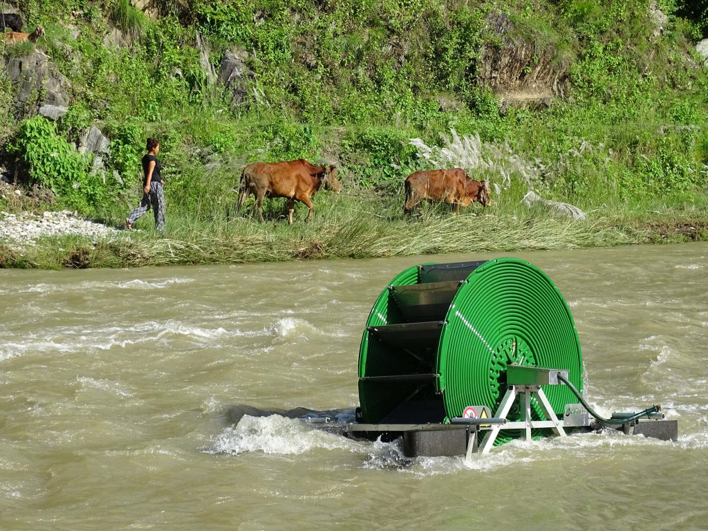 photo of water pump in river with woman and two cows walking on the far riverbank