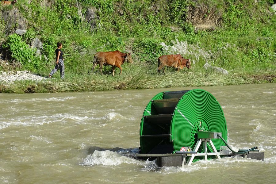 photo of water pump in river with woman and two cows walking on the far riverbank